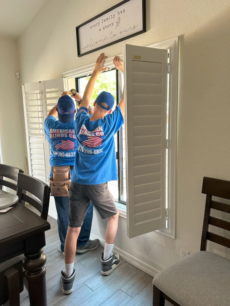 Two professional installers in blue shirts expertly fit energy-efficient white plantation shutters on a dining room window, enhancing comfort and style for homeowners. The elegant installation near a dining table and chairs promises increased energy savings, improved indoor comfort, and peace of mind with professional service.
