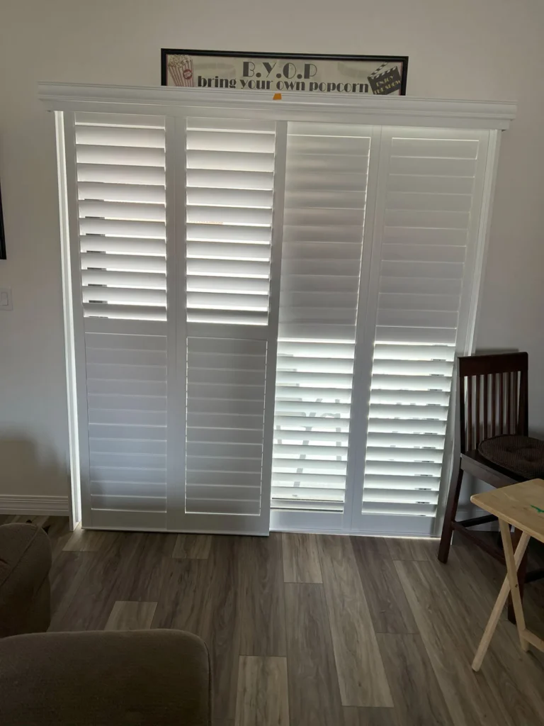 White plantation shutters, professionally installed on a large window, let in natural light while helping to lower energy costs and keep your space comfortable year-round. A cheerful "B.Y.O.P. bring your own popcorn." sign hangs above, with a cozy chair and small table nearby—creating an inviting, stylish room that’s both energy-efficient and relaxing.
