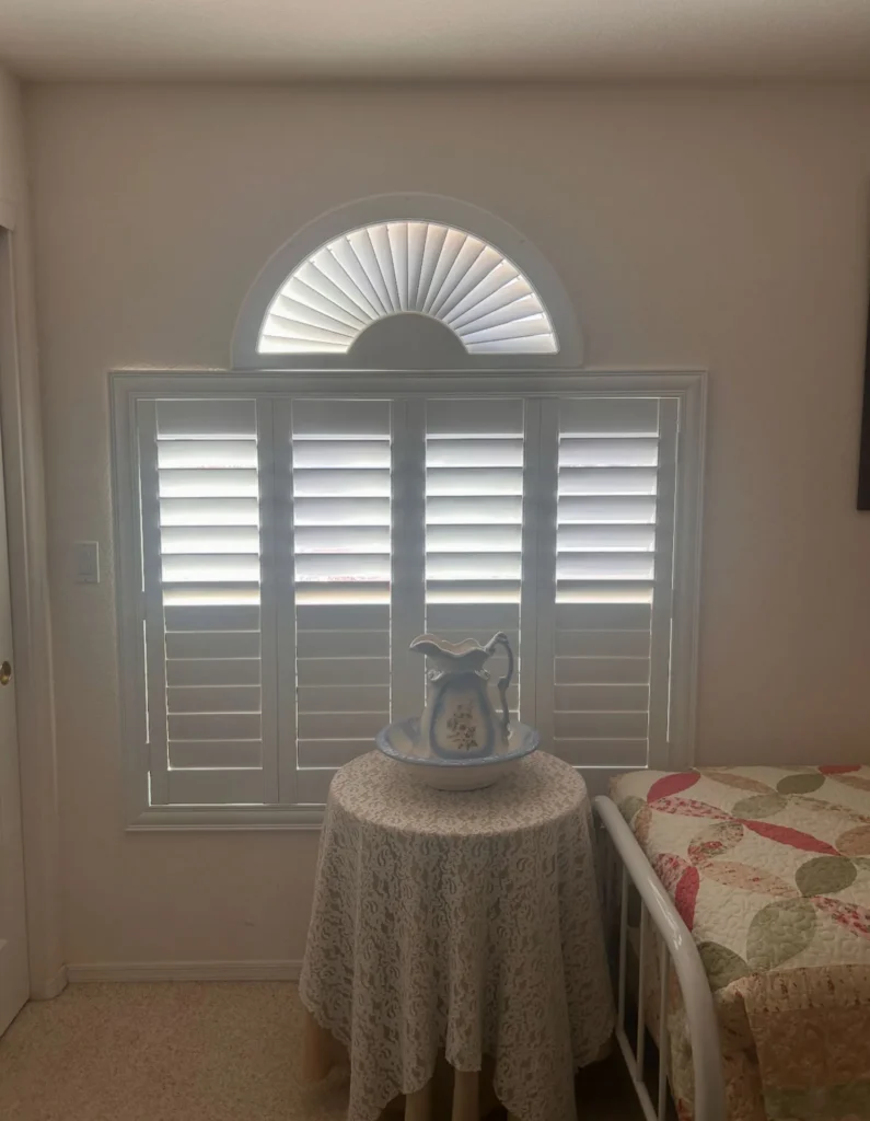 Image shows a bedroom window fitted with white plantation shutters and an arched fanlight above, offering increased energy savings and year-round comfort. Professional installation ensures a perfect fit next to the cozy bed with a patterned quilt, complemented by a round table adorned with an elegant lace cover and decorative pitcher.