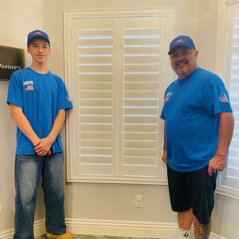 Two professional installers in matching blue uniforms stand indoors on either side of a window with closed white shutters, ready to provide expert installation for improved home comfort, increased energy savings, and a seamless upgrade experience.